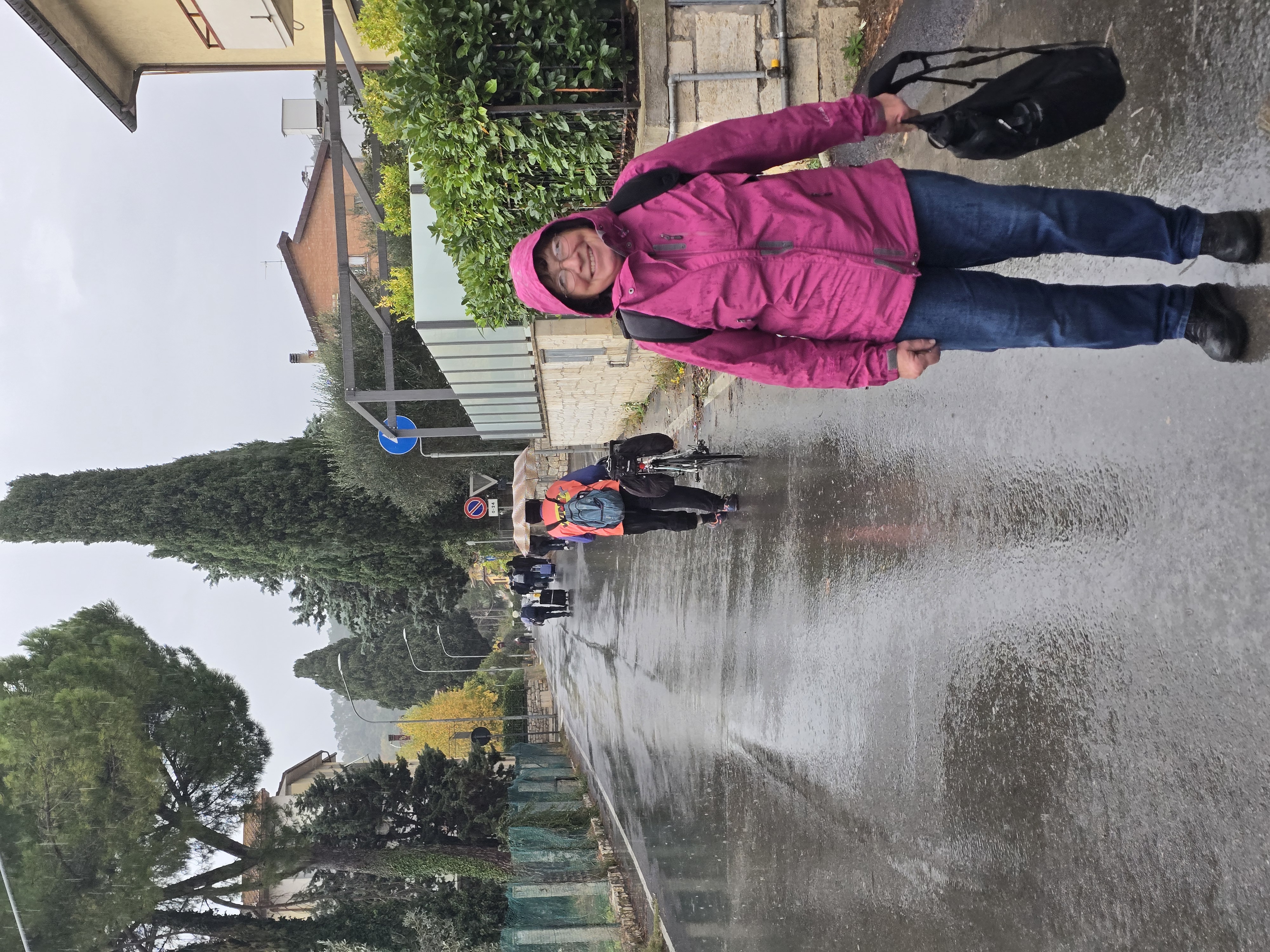 Rapolano Terme. Still in better spirits than me. My mom in a pink rain jacket, hood up and smiling, on a wet road at Rapolano Terme while the other stranded passengers walk on ahead